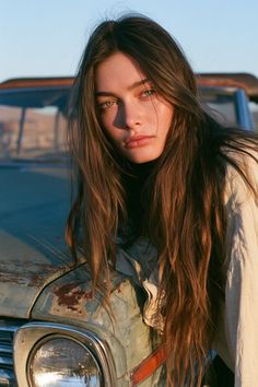Woman with long hair and green eyes leaning on an old car at golden hour