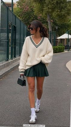 Woman in a cream v-neck sweater and green pleated skirt walking outdoors