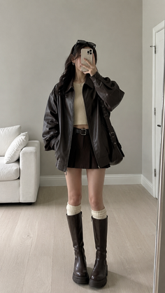 Person in a brown leather jacket and pleated skirt taking a mirror selfie