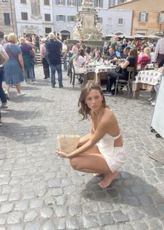 Woman in a white dress crouching on a cobblestone street with a small bag