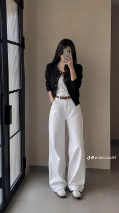 Woman in a black cardigan and white wide-leg pants taking a mirror selfie indoors