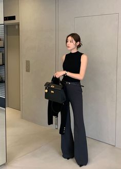 Woman in a black sleeveless top and flare pants holding a handbag indoors