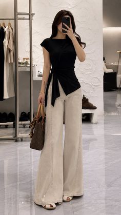Woman in a black top and wide leg pants taking a mirror selfie indoors