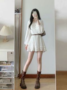 Woman in a white dress and brown boots taking a mirror selfie indoors