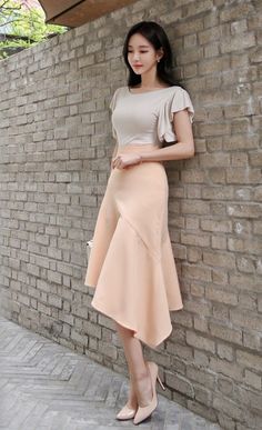 Woman in a cream top and peach midi skirt leaning against a brick wall