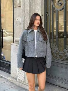 Woman with long hair wearing a grey jacket and black pleated skirt outdoors