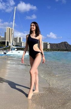 Woman in a black one-piece swimsuit walking on a beach with a cityscape in the background