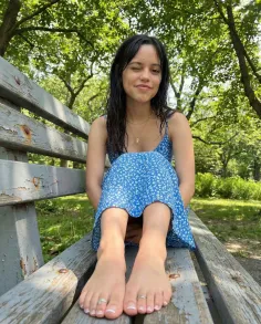 Woman in a blue floral dress winking while sitting barefoot on a park bench