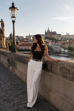 Woman in a black top and white pants leaning on a stone wall with a cityscape in the background