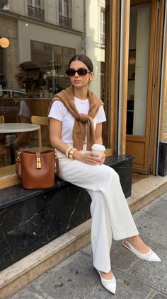 Woman in white pants and a brown sweater sitting outside a cafe with a coffee cup