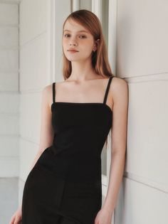Young woman with long hair wearing a black top leaning against a white wall