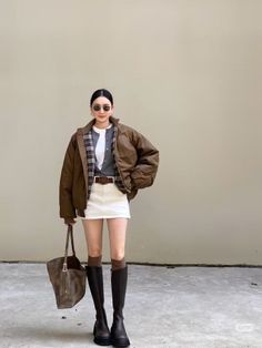 Woman in a brown jacket, white mini skirt, and knee high boots standing outdoors