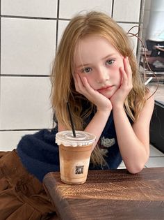 Young girl with long blonde hair resting her chin on her hands next to an iced coffee