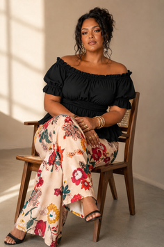 Woman with dark curly hair wearing an off-shoulder top and floral pants, sitting on a chair