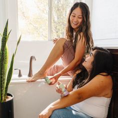 Two women in a bright bathroom, one sitting on the tub, holding small products