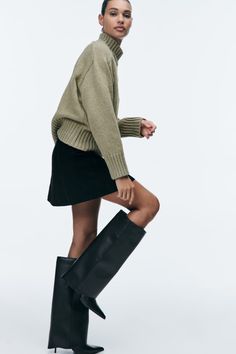 Woman in a turtleneck sweater, black mini skirt, and knee-high boots posing in a studio
