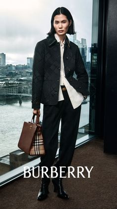 Woman in a quilted jacket and black pants holding a handbag indoors with a cityscape view
