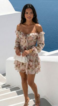Woman in a floral off-shoulder dress holding a white clutch outdoors by the ocean