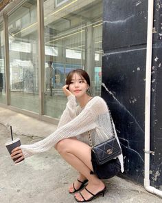 Woman in a sheer top and black shorts squatting outdoors with a drink
