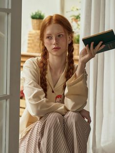 Woman with red hair and freckles wearing pajamas, holding a book indoors