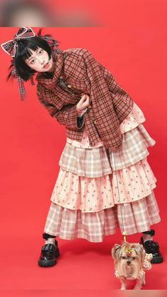 Person in a plaid jacket and tiered skirt with a small dog on a red background