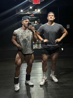 Two muscular men posing in a gym mirror, one taking a selfie