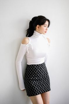 Woman with black hair in a white turtleneck and plaid skirt against a white wall
