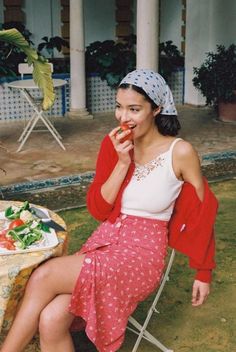 Woman in a polka dot scarf and red cardigan eating a tomato outdoors