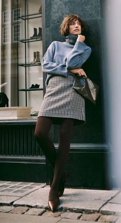 Woman in a blue turtleneck and plaid skirt leaning against a building