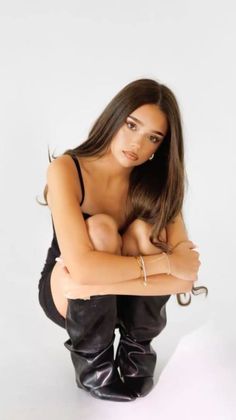 Woman with long brown hair in a black dress and boots squatting in a studio