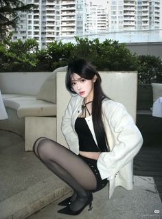 Woman with long black hair in a white jacket and sheer tights squatting outdoors
