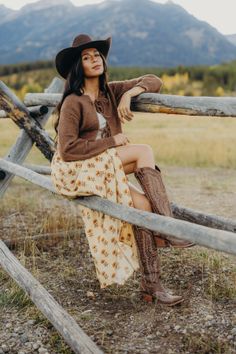 Woman in a cowboy hat and floral skirt sitting on a wooden fence outdoors
