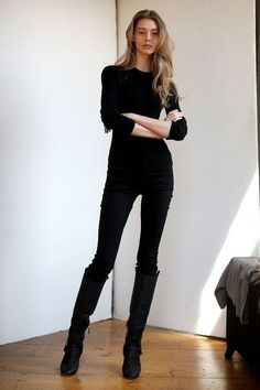 Woman with long hair wearing a black top, black pants, and black boots indoors