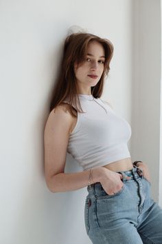 Woman with long hair in a white crop top and blue jeans leaning against a wall