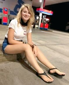 Blonde woman in a white t-shirt and denim shorts sitting at a gas station at night