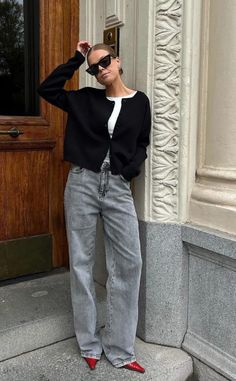 Woman in a black cardigan, grey jeans, and red shoes posing outdoors