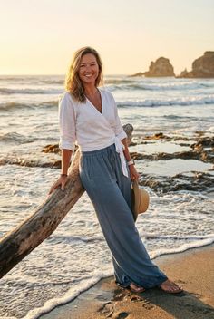 Woman in a white top and blue pants leaning on driftwood at the beach during golden hour