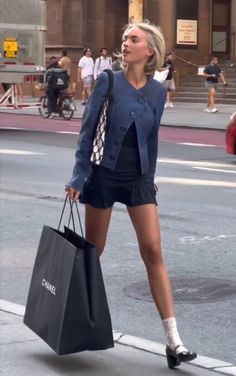 Woman with blonde hair in a blue jacket and black skirt walking on a street with a shopping bag