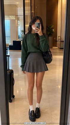 Woman in a green sweatshirt and pleated skirt taking a mirror selfie