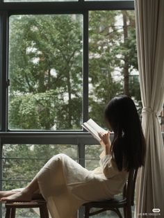 Woman with long dark hair reading a book by a window with a view of trees