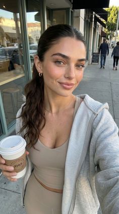 Woman in a beige crop top and leggings with a grey hoodie holding a coffee cup