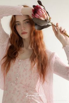 Woman with long red hair in a pink floral dress holding a protea flower