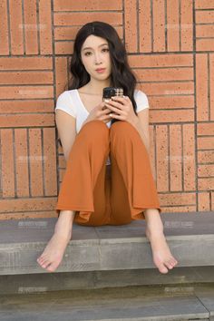 Woman with long dark hair sitting barefoot against a brick wall, holding a phone