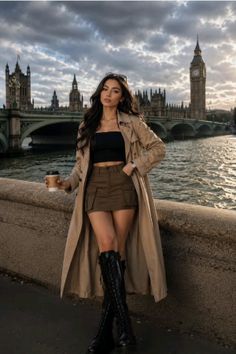 Woman in a trench coat and mini skirt holding coffee by a river with a cityscape