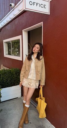 Woman in a tan jacket and lace shorts leaning against a red wall with a yellow tote bag
