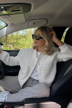 Woman with blonde hair and sunglasses in a white jacket and striped pants inside a car