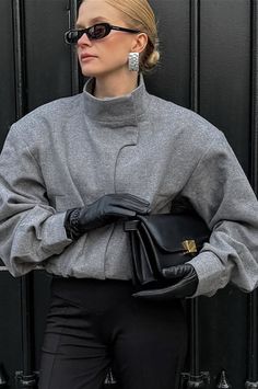 Woman in a gray jacket, black gloves, and sunglasses holding a black handbag