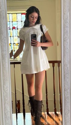 Woman in a white dress and lace-up boots taking a mirror selfie indoors