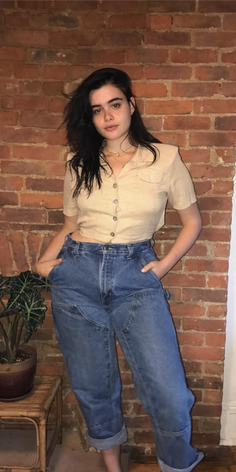 Woman with dark hair wearing a cropped shirt and baggy jeans against a brick wall