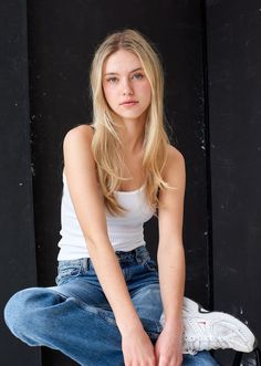 Young woman with blonde hair wearing a white tank top and blue jeans against a dark background